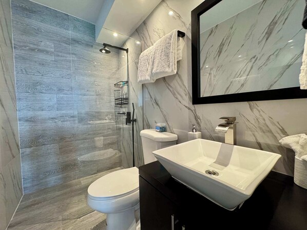 A modern bathroom with a glass-enclosed shower, a white toilet, a large vessel sink, and a marble backsplash.