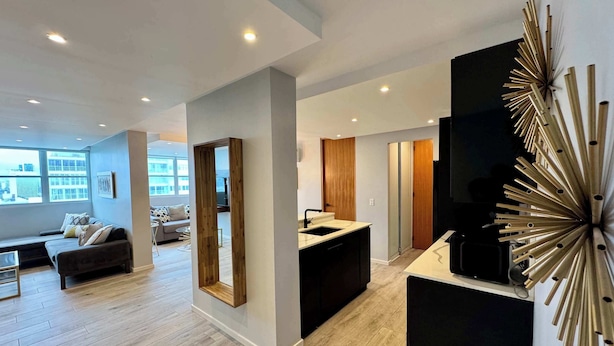 Una sala de estar moderna con sofá, una mesa de centro y un espejo grande. Cuenta con una zona de cocina con un fregadero y un refrigerador. El cuarto tiene piso de madera y un ventanal amplio.