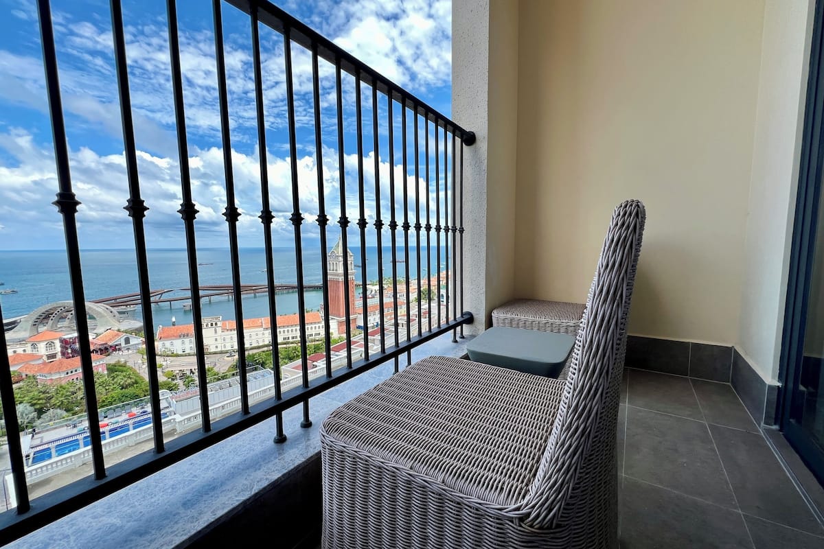 A balcony with a wicker sofa and a small table, overlooking a coastal cityscape with a prominent tower and a bridge.