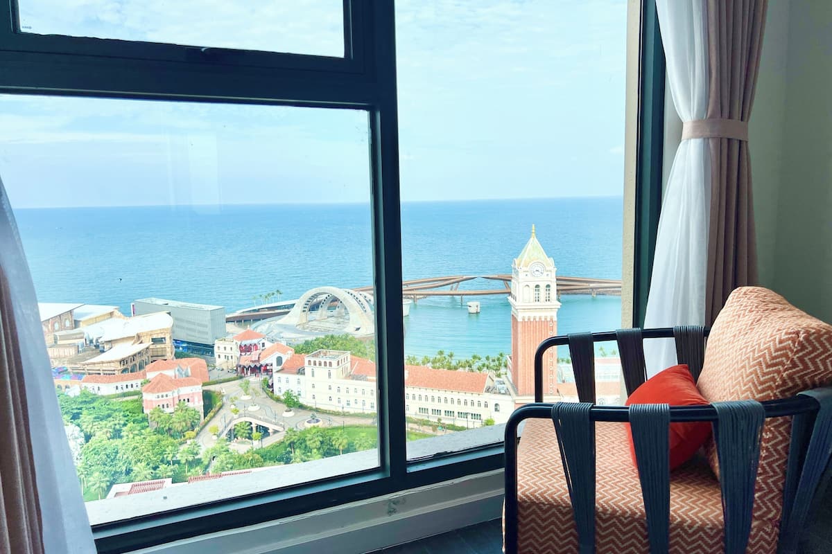 A room with a large window overlooking a coastal cityscape, featuring a clock tower and a bridge.