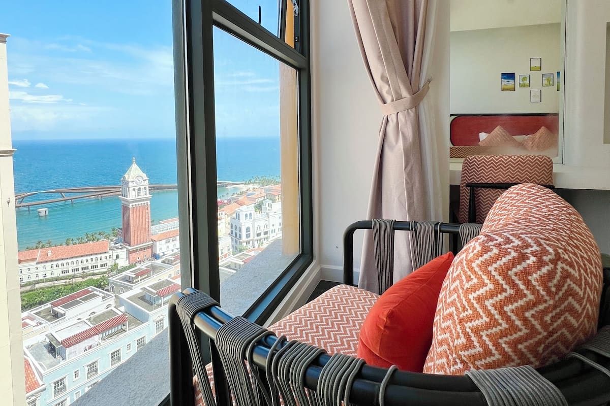A room with a view of a coastal cityscape, a bed with patterned pillows, and a window with curtains.