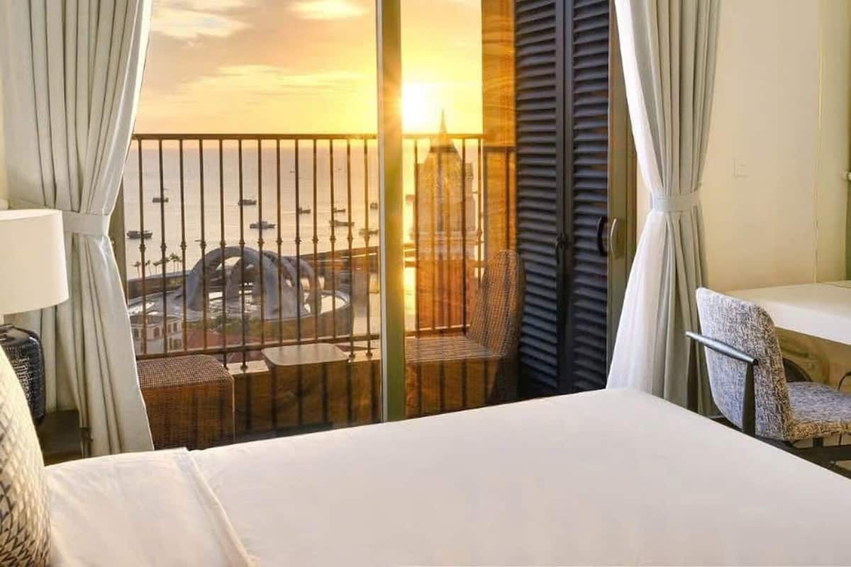 A hotel room with a bed, a chair, and a balcony with a view of a cityscape and sunset.