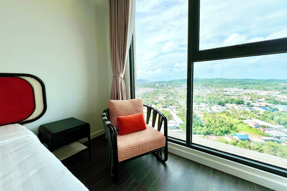 A hotel room with a large window offering a city view, a bed, a chair with a red pillow, and a small table.