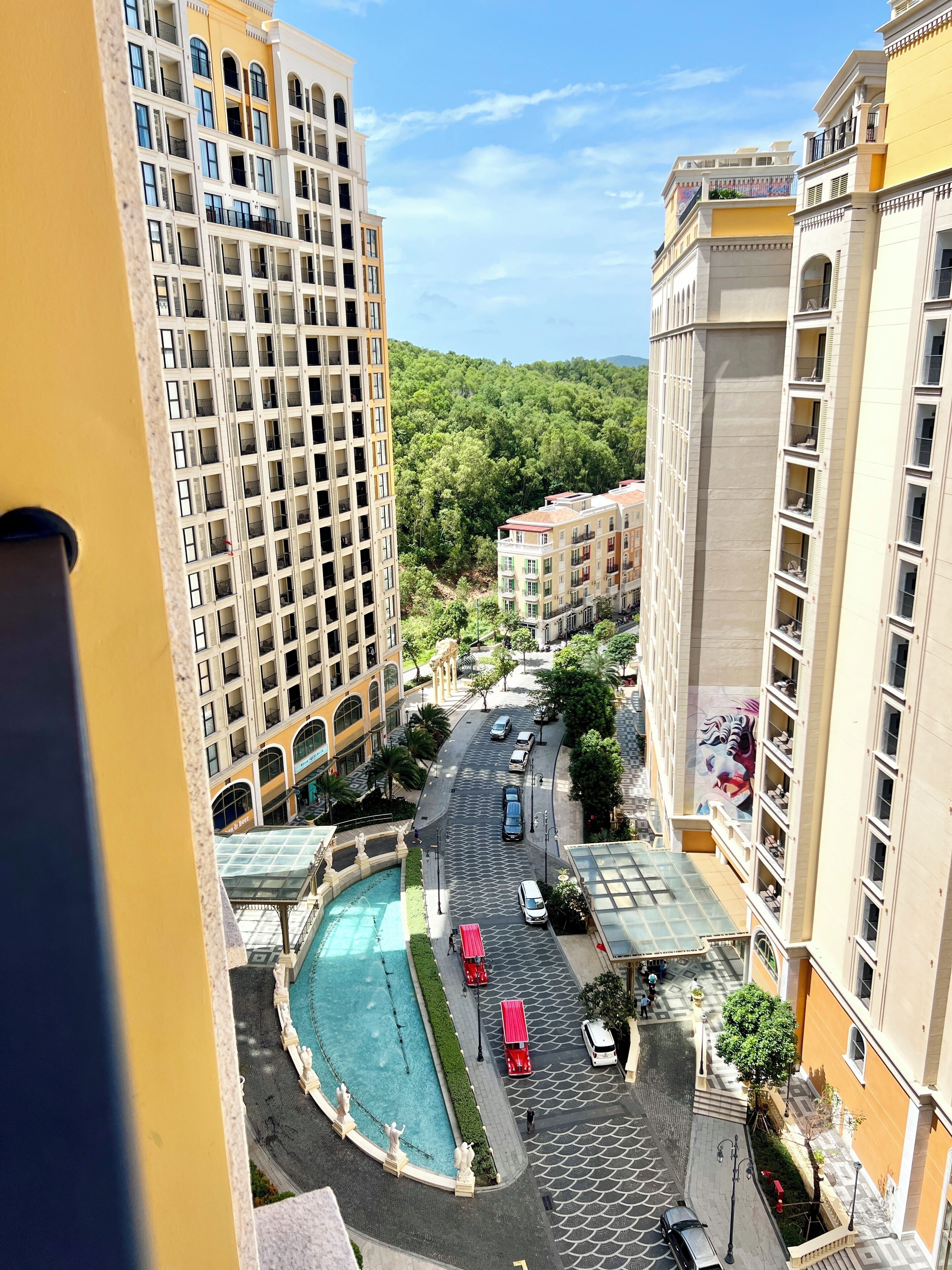 Un hotel moderno de gran altura con varios balcones, un patio central con una fuente y zonas verdes y coches aparcados en los alrededores.