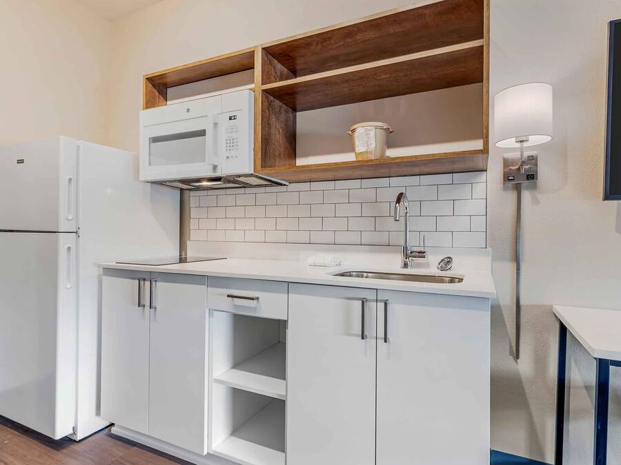 A compact kitchen with white cabinetry, a built-in microwave, a sink, and a wooden shelf above the countertop.