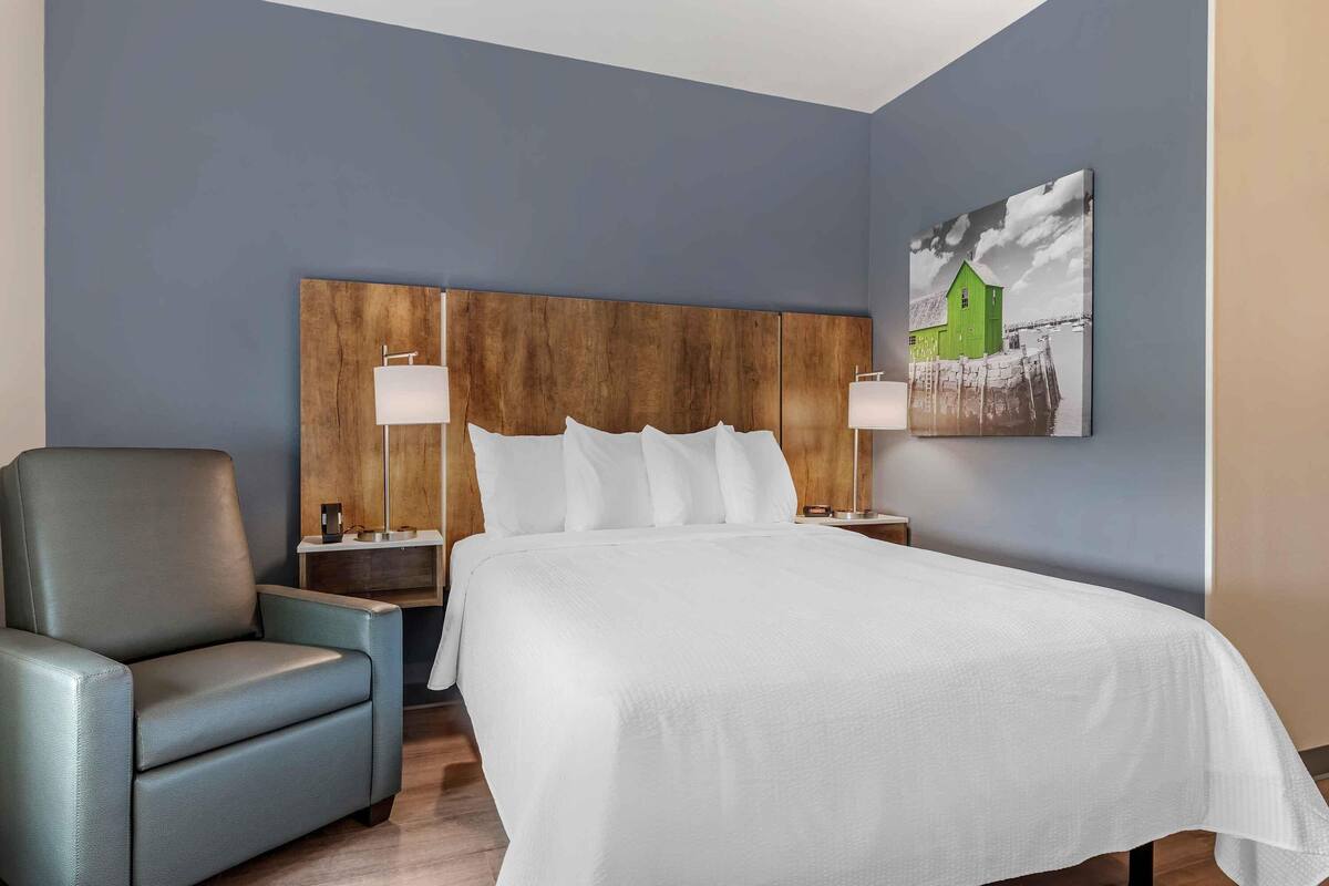 A hotel room with a large bed, a wooden headboard, a blue armchair, and a painting of a green house on the wall.