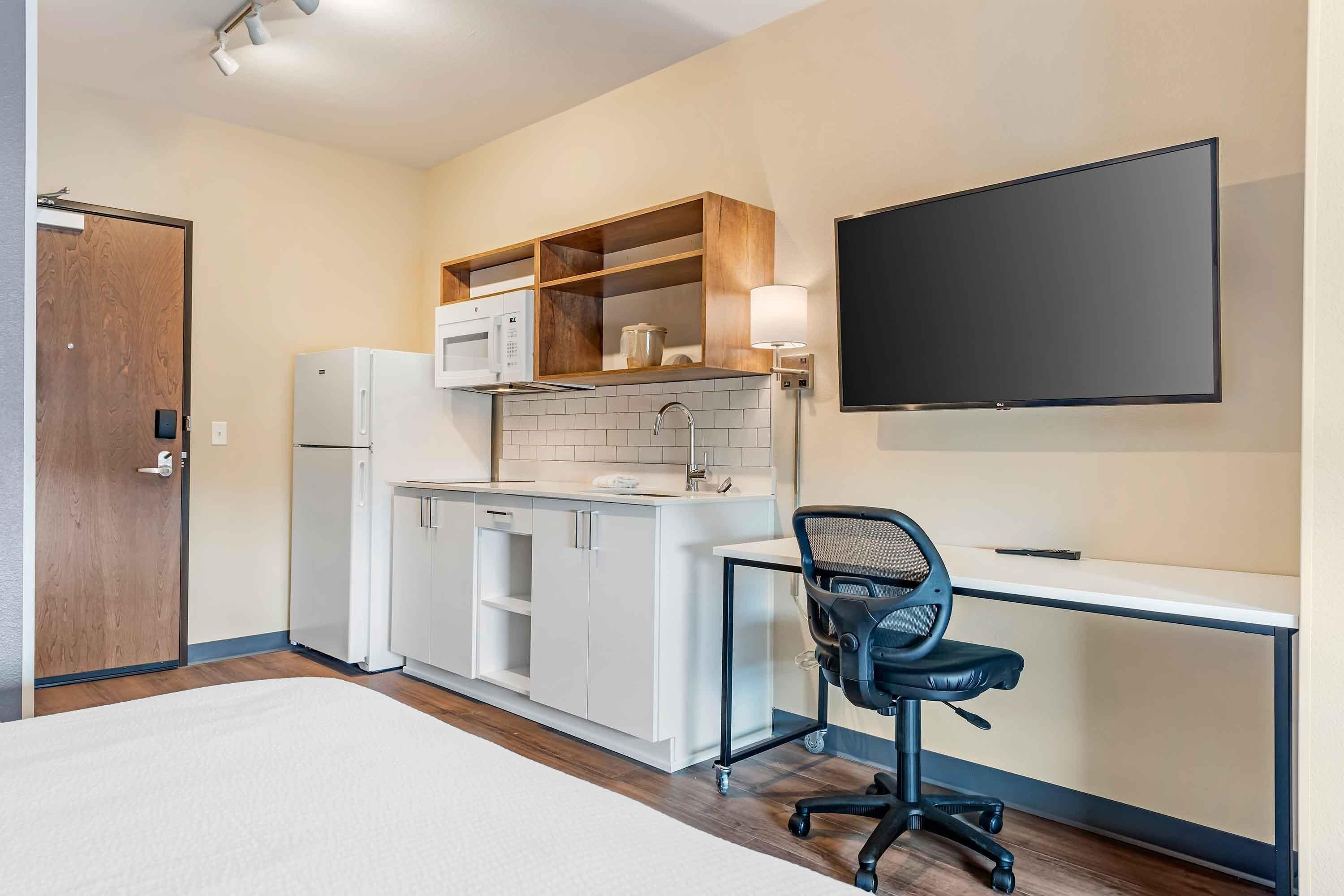 A compact hotel room with a kitchenette, a bed, a desk with a chair, and a TV mounted on the wall.