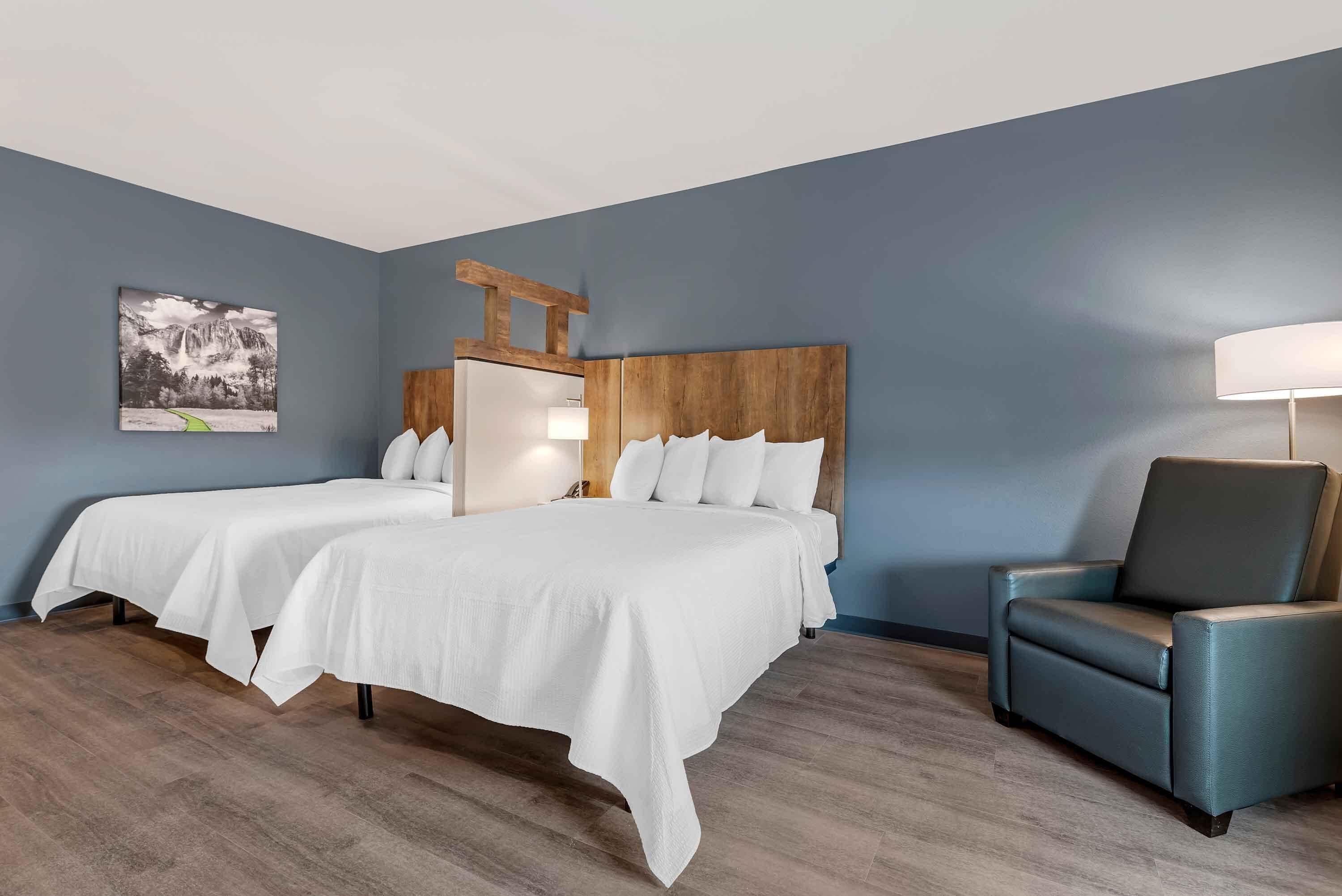 A hotel room with two beds, a wooden headboard, a blue armchair, and a wall-mounted lamp.