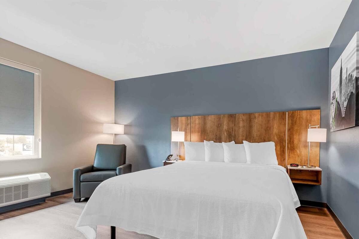 A hotel room with a large bed, a chair, a nightstand, a lamp, and a wall-mounted picture.