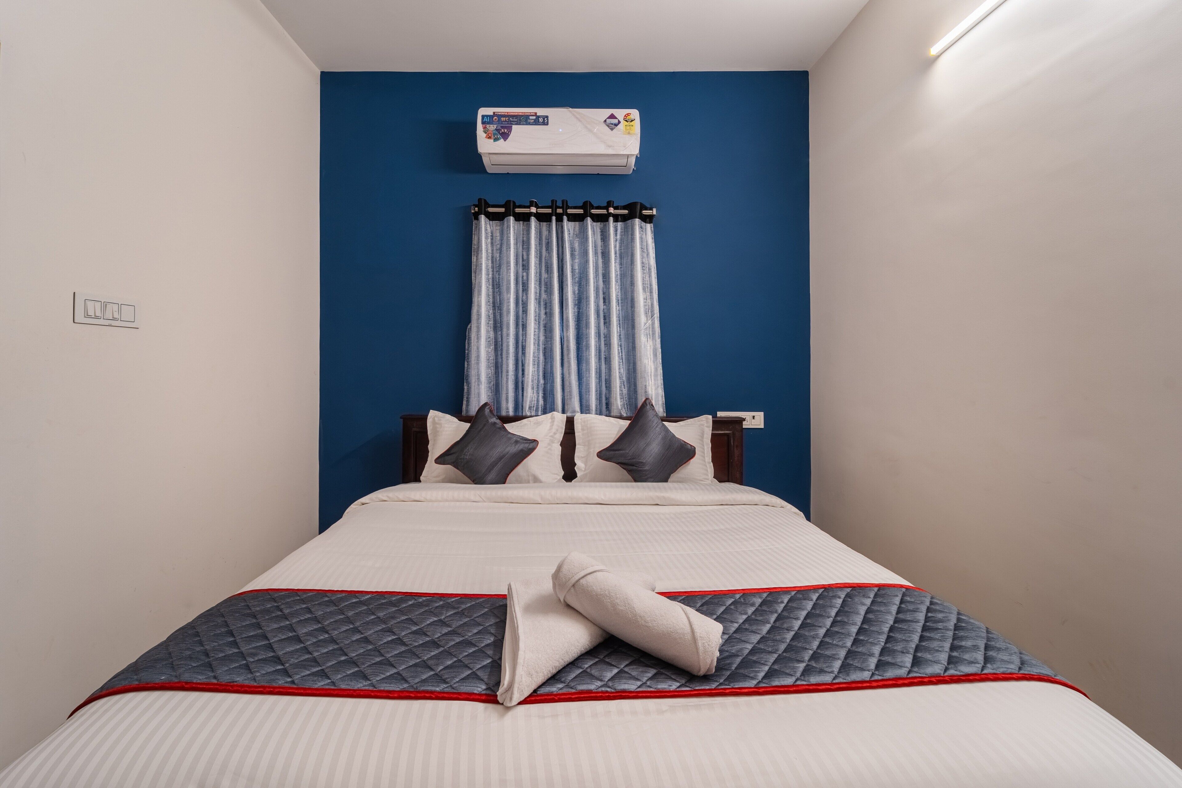 A neatly made bed with a quilted blanket, a wall-mounted air conditioner, and a curtain.