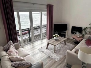 A living room with a sofa, coffee table, TV, and a view of the sea through large glass doors. - Sea front Apartment in Tramore (Tramore)