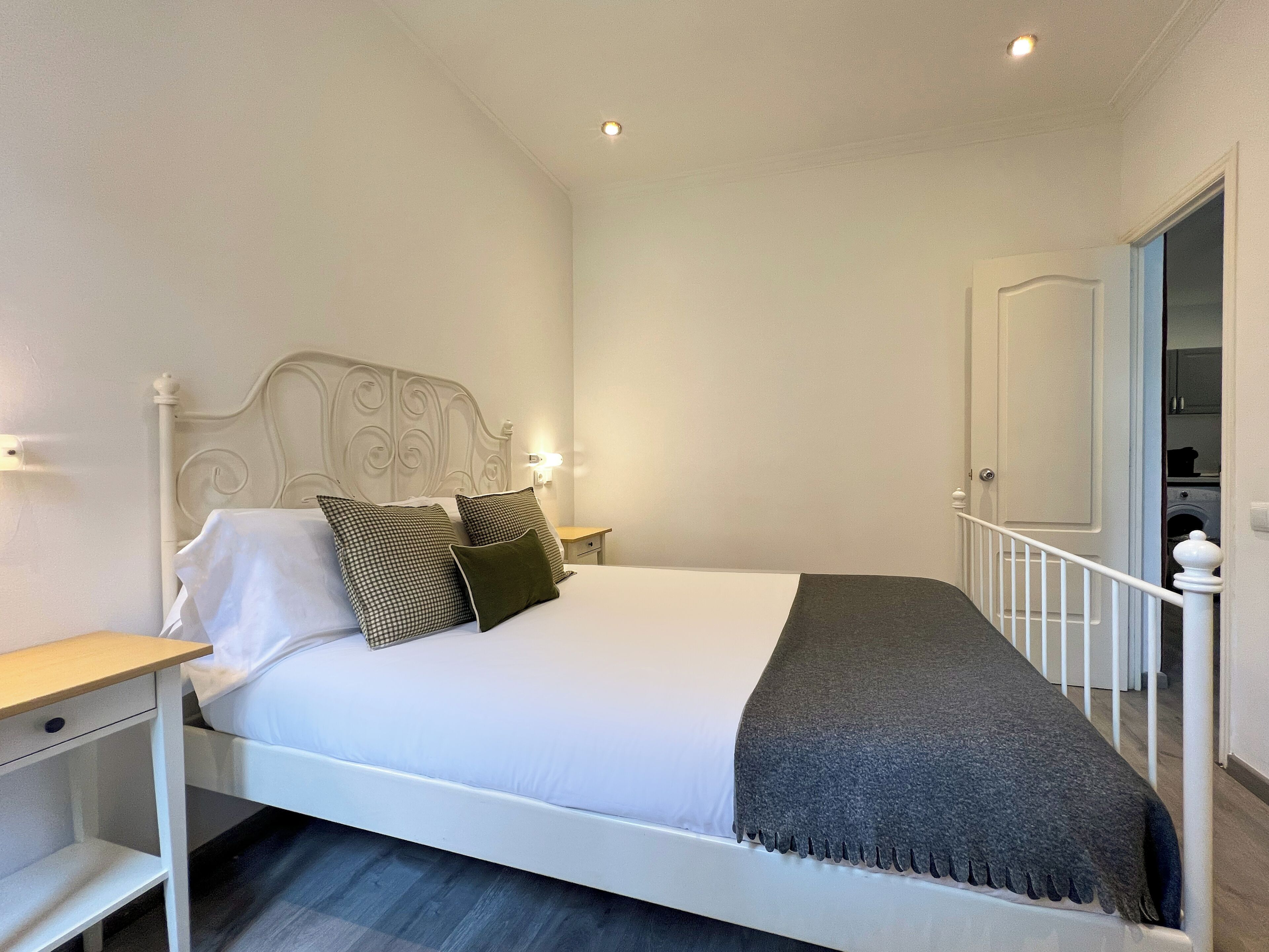 A neatly made bed with white bedding and two pillows, a bedside table with a lamp, and a door leading to a laundry area.