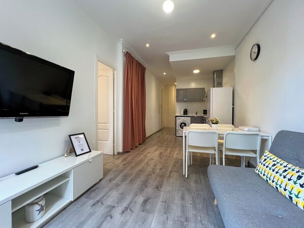 A modern apartment with a flat-screen TV, a dining area, a sofa, and a kitchen in the background.