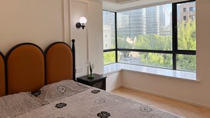 A bedroom with a large window, a bed with a headboard, a nightstand, and a wall-mounted light fixture.
