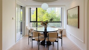 A dining area with a round table, six chairs, a vase with flowers, and a large window with a city view.