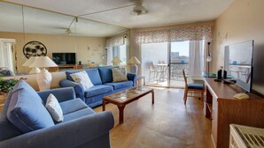 A living room with a blue sofa, a coffee table, a TV, and a balcony with a view. - 254 El Matador 1 Bedroom Condo (Fort Walton Beach)