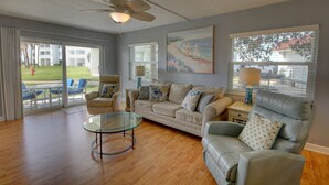 A spacious living room with a beige sofa, a glass coffee table, and a blue armchair. There is a ceiling fan, a painting on the wall, and a view of the outside through large windows. - 119 El Matador 2 Bedroom Condo (Fort Walton Beach)