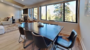 A modern living room with a large dining table, comfortable seating, and large windows offering a view of trees. - 1849 Condos 642 3 Bedroom Home (Mammoth Lakes)