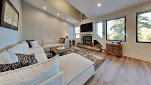 A modern living room with a sofa, a coffee table, a fireplace, and large windows. - 1849 Condos 642 3 Bedroom Home (Mammoth Lakes)
