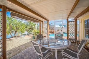 Un porche cubierto con una mesa y sillas de vidrio, con vista a una piscina y palmeras.