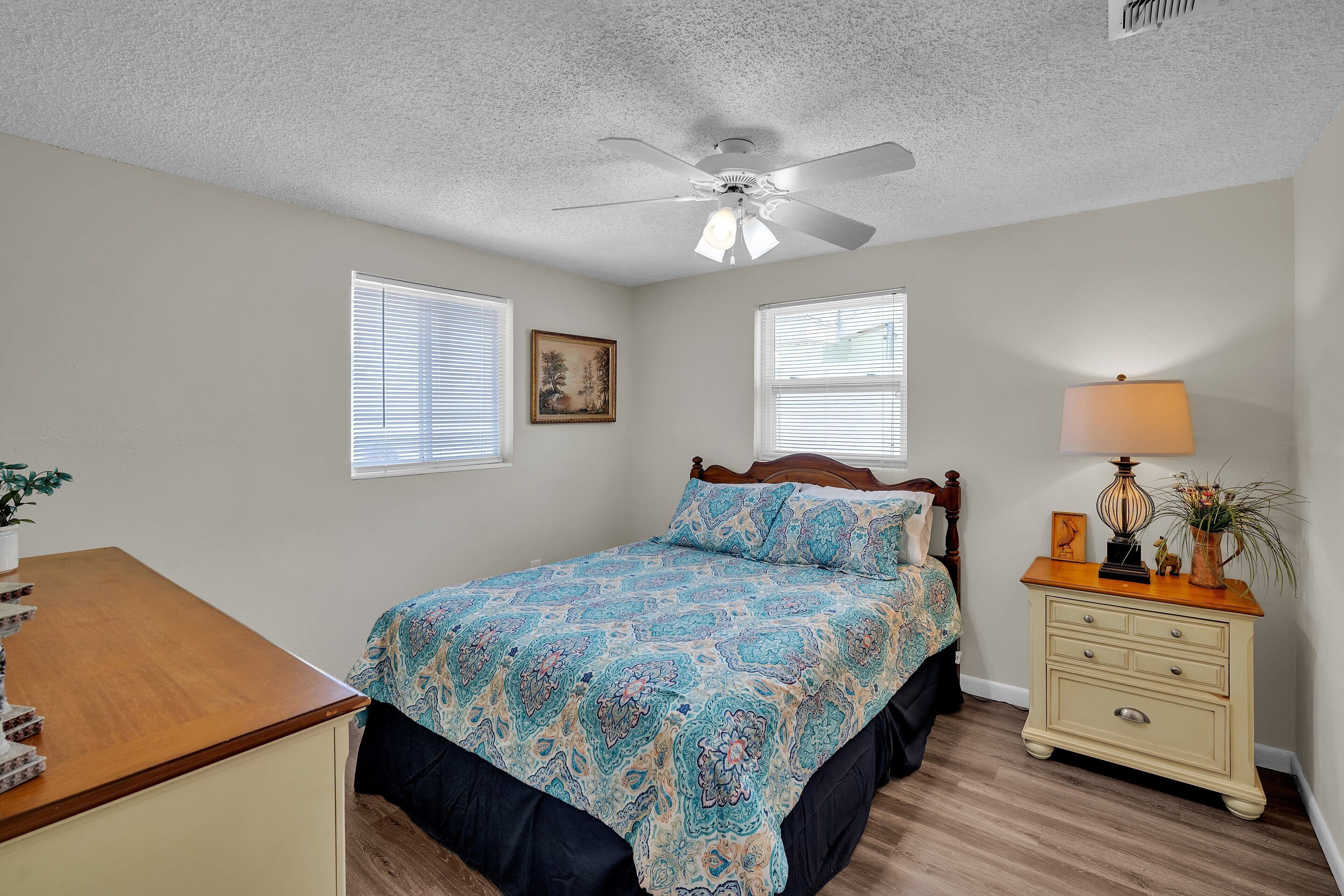 A bedroom with a bed, a nightstand, a ceiling fan, and two windows with blinds.