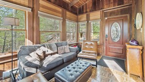A cozy room with a large window, a wooden ceiling, a sofa, a chair, and a door with a glass panel. - Charming Porter House In The Heart Of The Poconos 4 Bedroom Chalet (Bushkill)