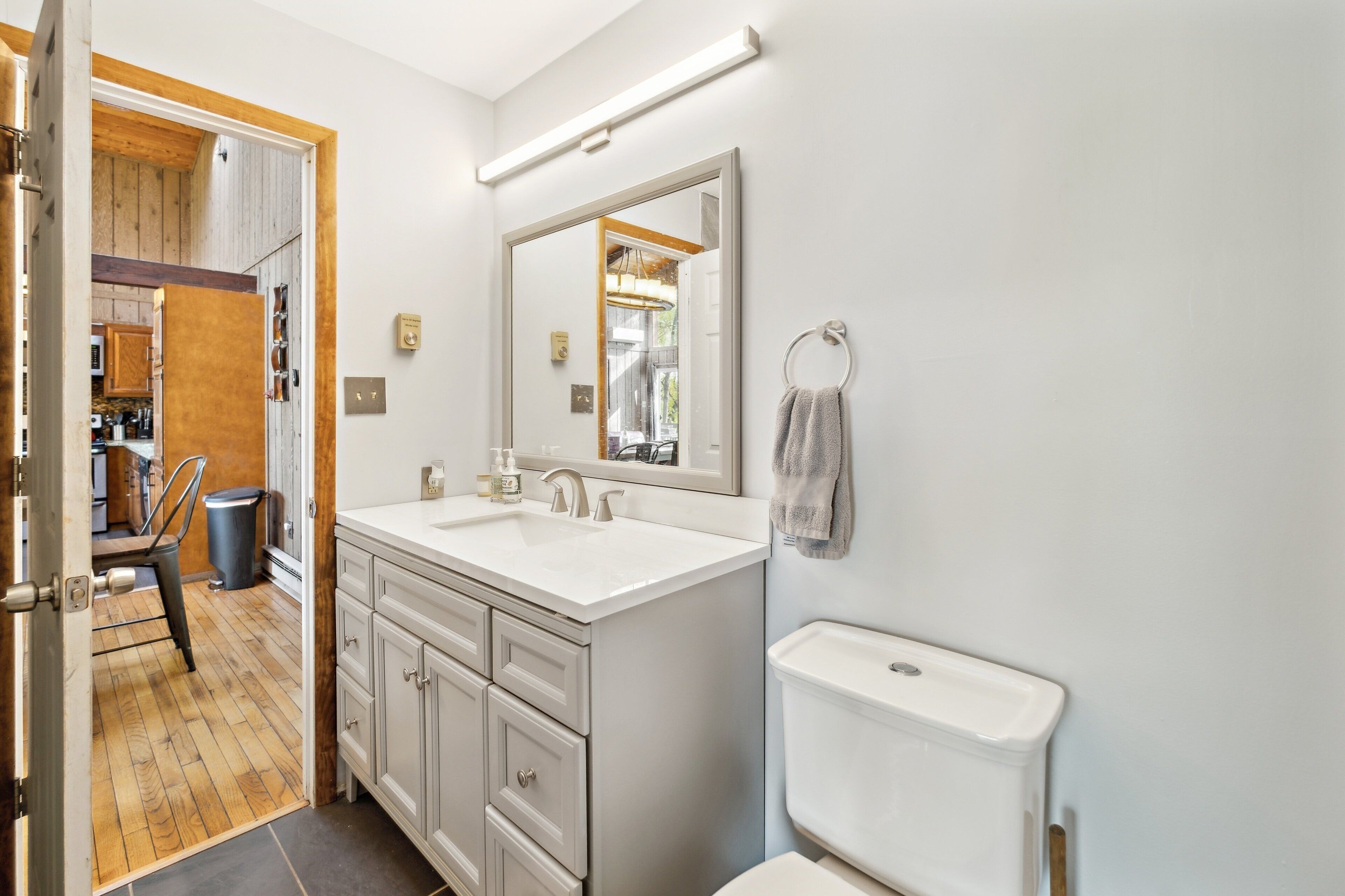 A modern bathroom with a large mirror, white sink, and toilet. There is a towel ring on the wall and a trash can in the corner.