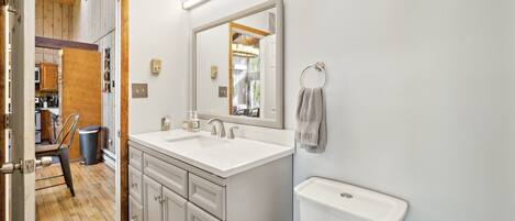 A modern bathroom with a large mirror, white sink, and toilet. There is a towel ring on the wall and a trash can in the corner.