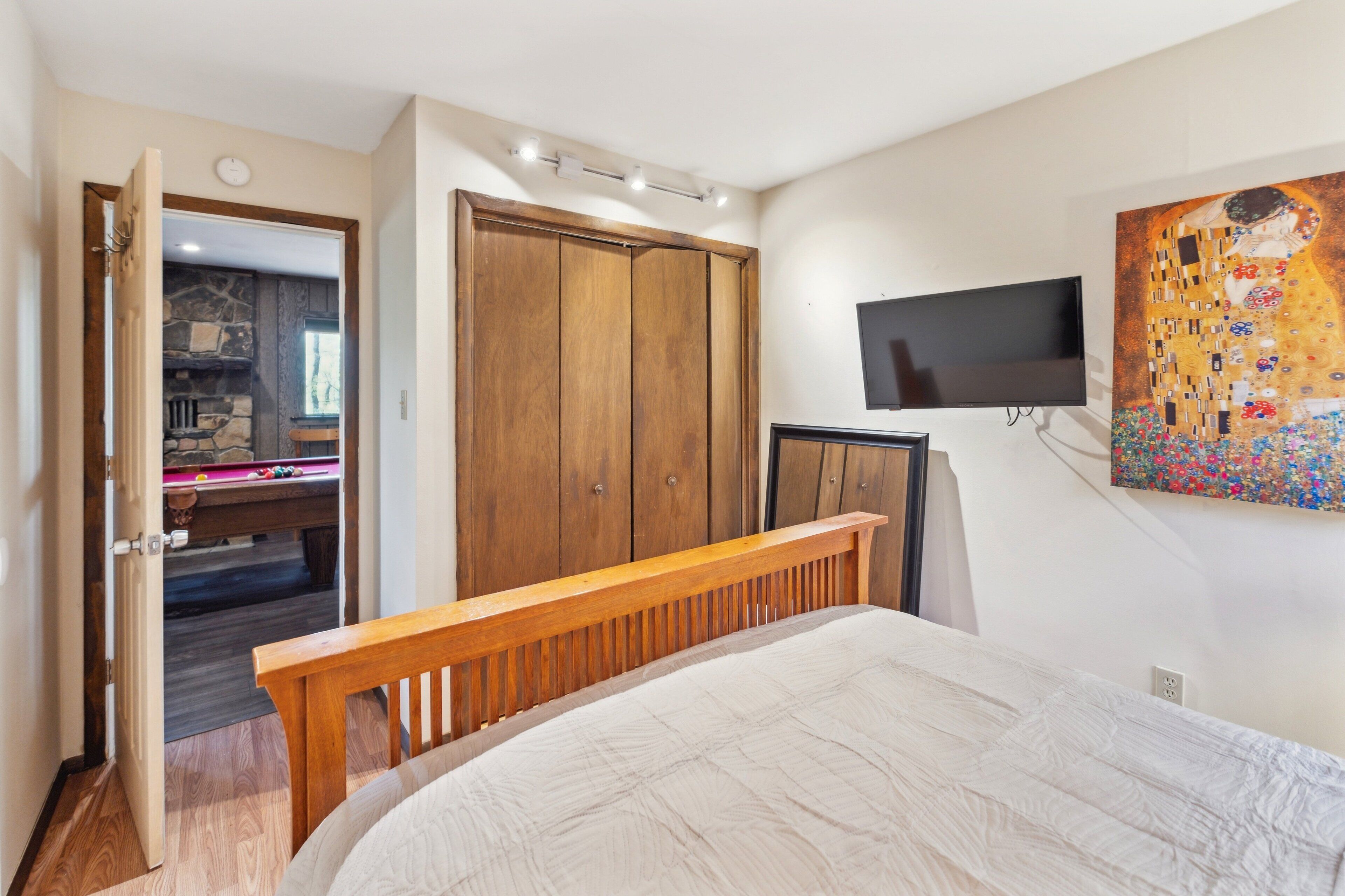A bedroom with a wooden bed, a TV mounted on the wall, a wooden wardrobe, and a painting.
