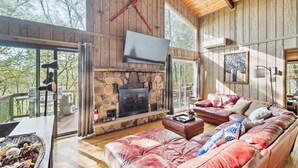 A cozy living room with a stone fireplace, leather sofa, and a large window overlooking a deck. - Charming Porter House In The Heart Of The Poconos 4 Bedroom Chalet (Bushkill)