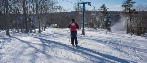 A person in a red jacket standing on a snow-covered ski slope with ski lifts and trees in the background.