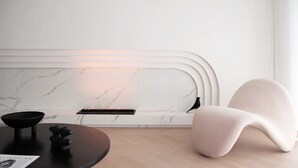A modern, minimalist room with a curved white headboard, a round wooden table, and a plush, light-colored armchair.
