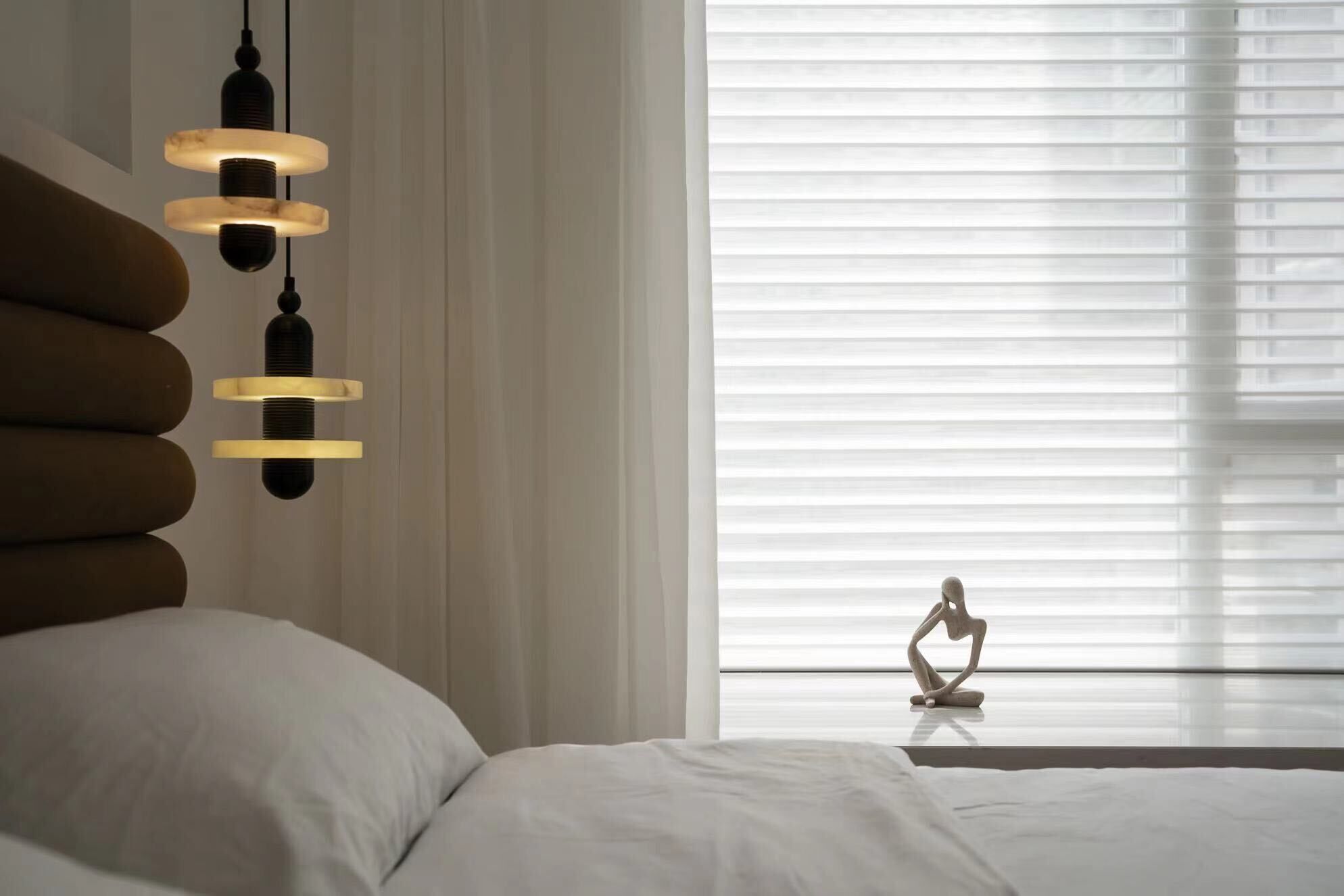 A bedroom with a bed, a hanging light fixture, a window with blinds, and a sculpture on the windowsill.