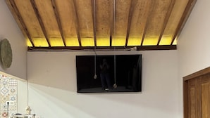 A room with a wooden ceiling, a ceiling fan, a wall-mounted TV, a wooden bench, and a patterned floor. - Omah Ndoro Sugih (Triwidadi)