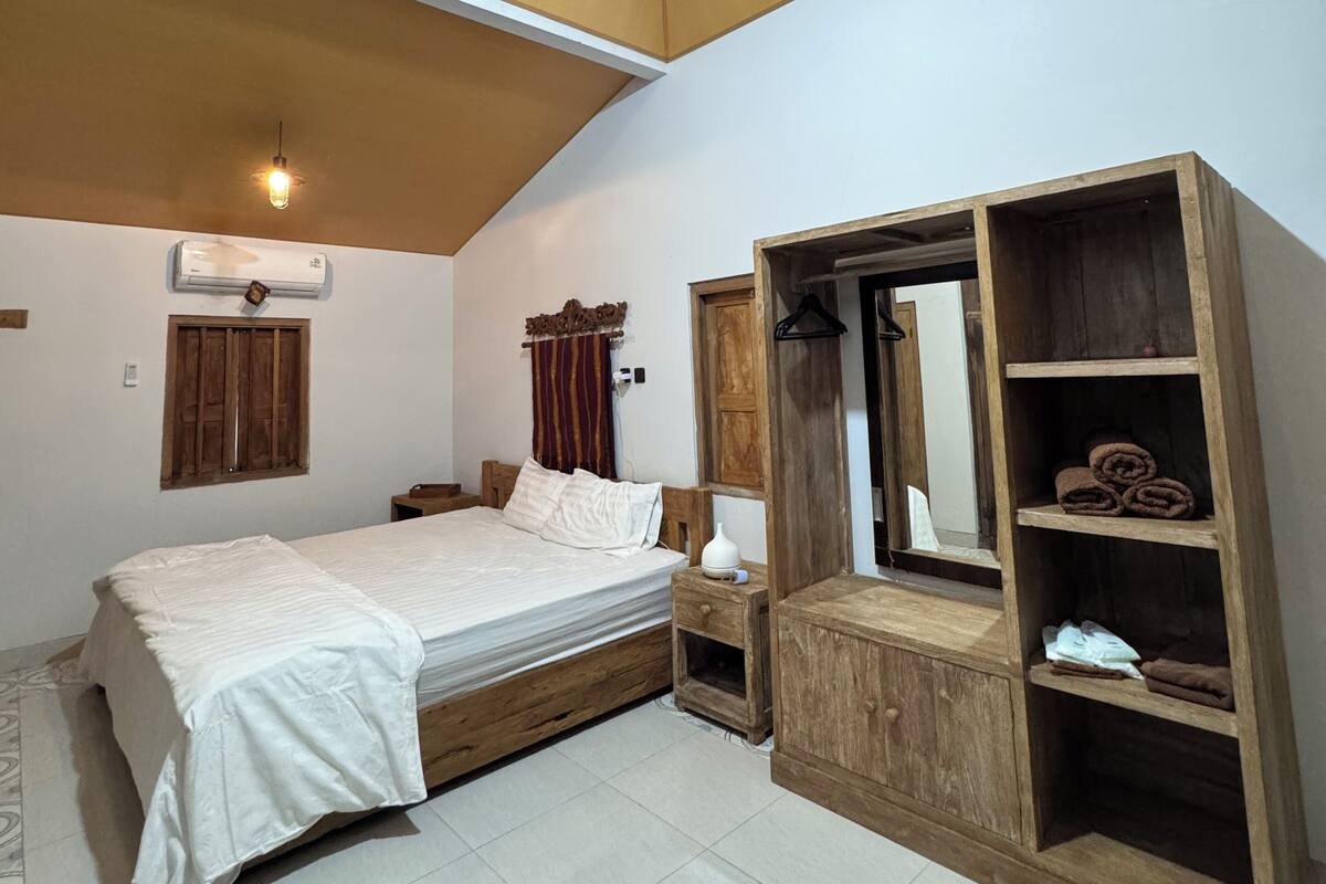 A bedroom with a wooden ceiling, a bed with a white bedspread, a wooden wardrobe, a bedside table, and a wall-mounted air conditioner.