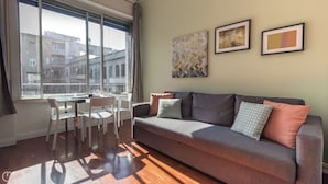 Un soggiorno con un divano grigio, cuscini decorati, un tavolino da caffè in vetro e sedie bianche. Sono presenti due opere d'arte incorniciate sulla parete e una finestra con vista sugli edifici.