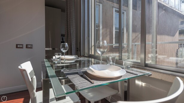 Zona pranzo con tavolo in vetro, sedie bianche e vista su un edificio in mattoni attraverso ampie finestre.