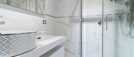 Un baño moderno con cabina de ducha de mármol, lavamanos blanco con jabón estampado y un espejo grande.
