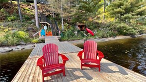 Eine Holzsteg mit zwei roten Adirondack-Stühlen, die auf einen Fluss ausgerichtet sind. Im Hintergrund ist eine kleine Holzkonstruktion mit einem roten Kanu zu sehen.