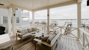 A covered outdoor deck with wooden flooring, white railings, and multiple seating areas including sofas and tables. - The Seafarer (Wrightsville Beach)