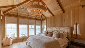 A wooden cabin bedroom with a large bed, a hanging light fixture, and a view of the water through large windows. - The Seafarer (Wrightsville Beach)