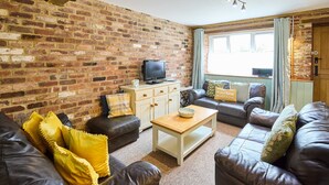 A living room with a brick wall, leather sofas, a wooden coffee table, and a television. - Host Stay West Barn (Fakenham)