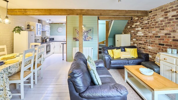 A cozy living area with a black leather sofa, wooden coffee table, and a dining table with chairs. A kitchen is visible in the background with a brick wall and wooden beams. - Host Stay West Barn (Fakenham)