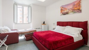 Une chambre d’hôtel moderne avec un grand lit, un canapé rouge, une petite table et une fenêtre donnant sur des immeubles.