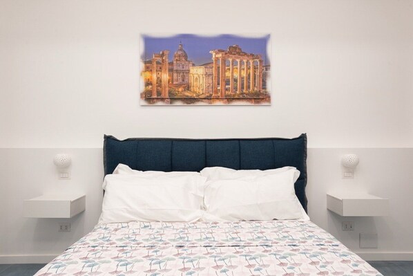 Une chambre à coucher avec un lit, des oreillers blancs, une tête de lit bleue, un tableau représentant des ruines antiques accroché au mur, et des tables de chevet avec des lampes.
