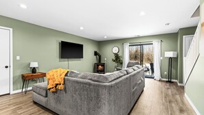 A modern living room with a grey sectional sofa, a flat-screen TV mounted on the wall, a small table with a lamp, and a fireplace. - Northern Comfort: The Vault - Your Refined Retreat near Duluth (Hermantown)