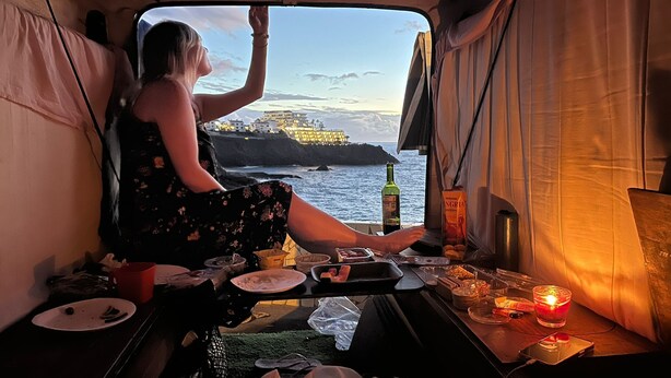 A person sitting by a window in a vehicle, with a bottle and snacks on the table. - Vintage Camper Van in Mallorca, with everything for live, 9 persons driving. (Sol de Mallorca)