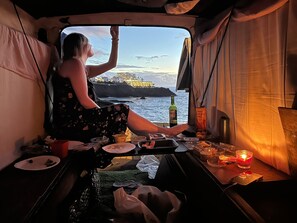 A person sitting by a window in a vehicle, with a bottle and snacks on the table. - Vintage camper van in Mallorca, fully equipped for living, seats 9 (Sol de Mallorca)