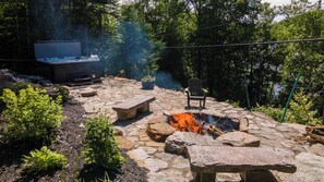 A stone patio with a fire pit, a hot tub, and a chair. - Lake | Pool Table | Au Pied du Lac (Labelle)