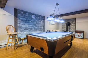 A pool table in a room with a wooden floor, a chair, a clock on the wall, and a hanging light fixture. - Lake | Pool Table | Au Pied du Lac (Labelle)